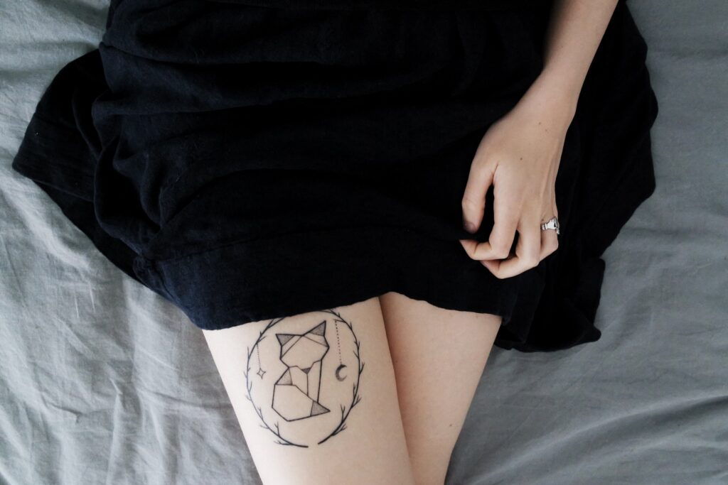 Thighs of a woman with a short black dress. Her right thigh has a cat tattoo.